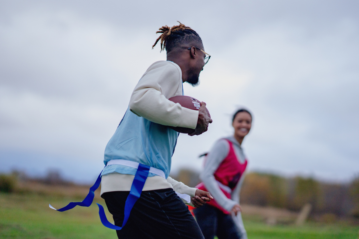 A group of University students run around on a field as they play a game of flag football together.  They are each dressed comfortably and have numbered team pinnies on.